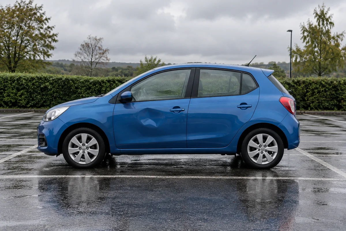 Voiture citadine bleue vue de profil sur un parking désert, portière avant légèrement enfoncée, sous un ciel couvert