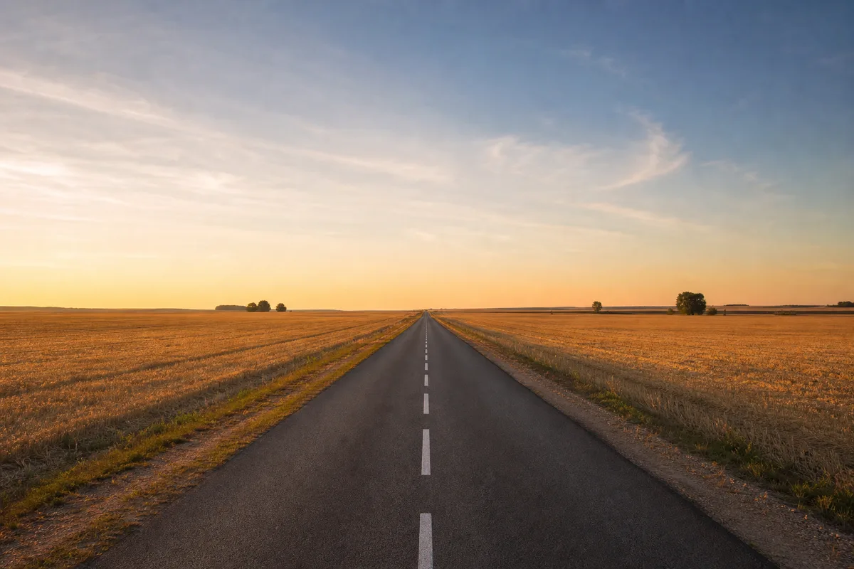 Route départementale rectiligne bordée de champs moissonnés sous une lumière dorée de fin de journée