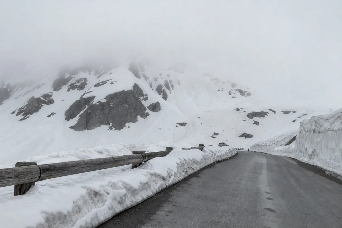 Col alpin désert sous un ciel bas, congères de neige damée bordant la chaussée et barrières de bois