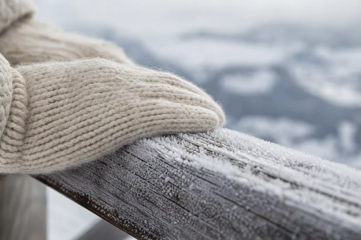 Mains gantées de laine posées sur une rambarde en bois couverte de givre, vallée en contrebas