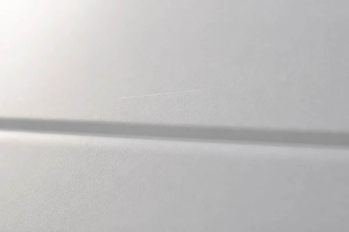 Tôle blanche laquée en gros plan, nervure horizontale et fine rayure sous une lumière rasante