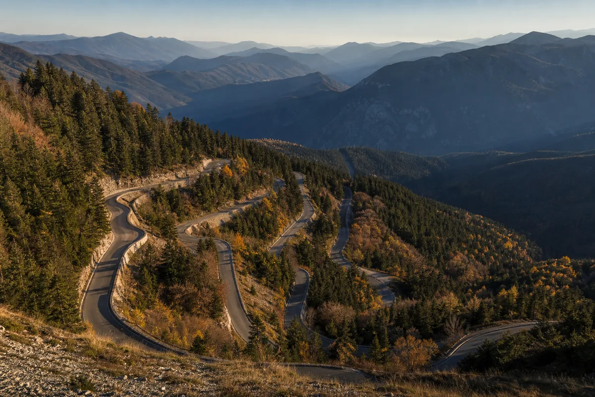 Route de montagne en lacets serrés vue depuis un belvédère boisé sous une lumière rasante