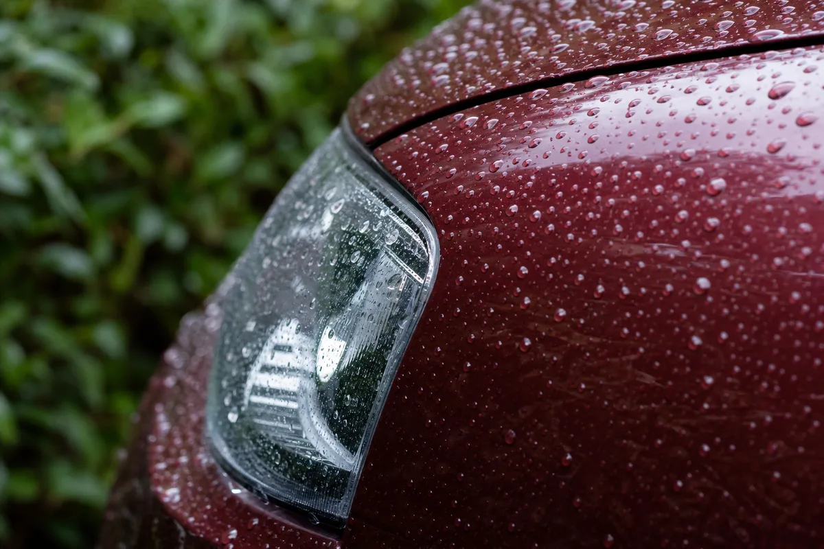 Détail d’un phare avant et d’une aile de voiture couverts de gouttes de pluie, végétation floue à l’arrière-plan